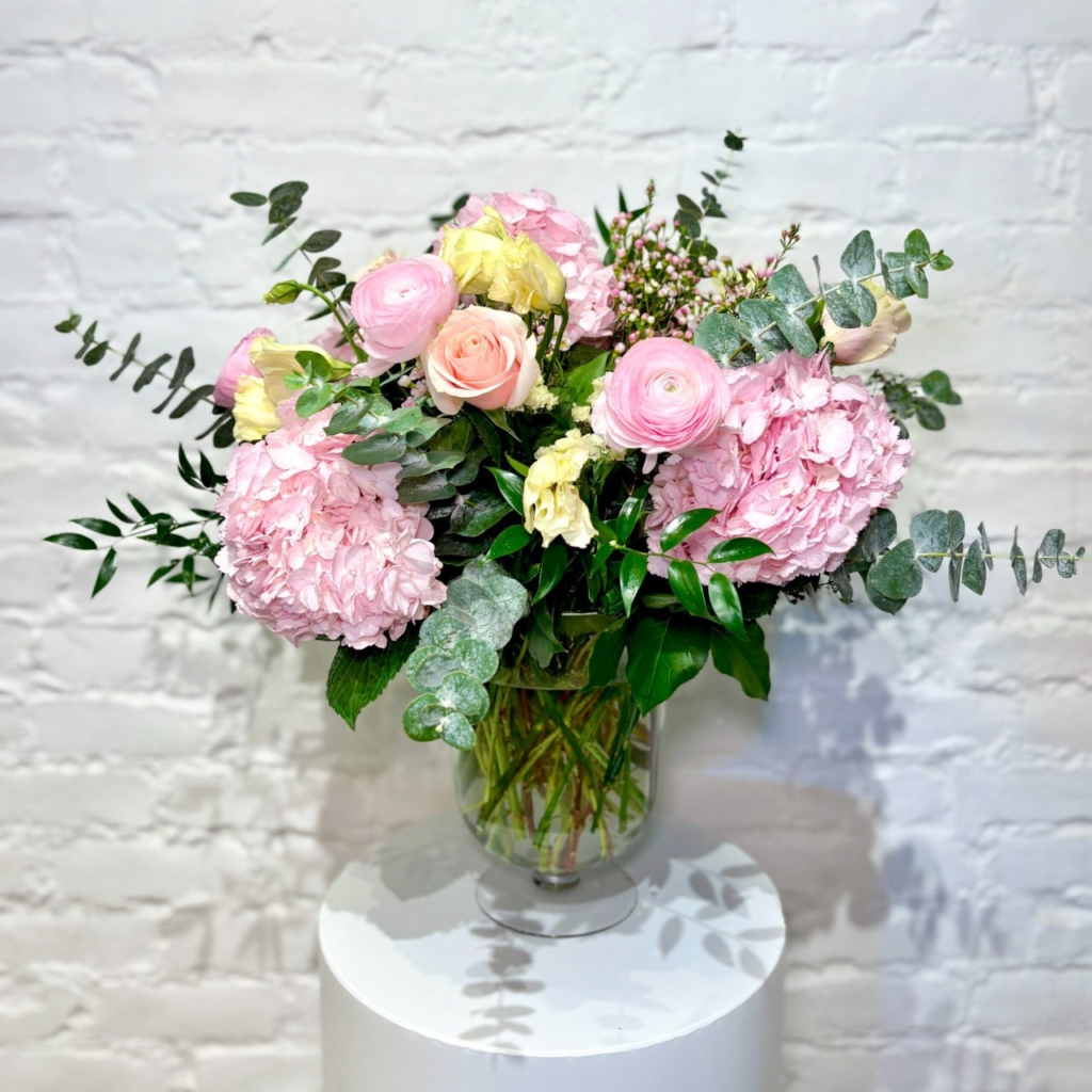 Fluffy pink hydrangeas, beautiful roses, show-stopper ranunculus, lisianthus and wax flower, expertly arranged with lush greens in a clear glass vase.