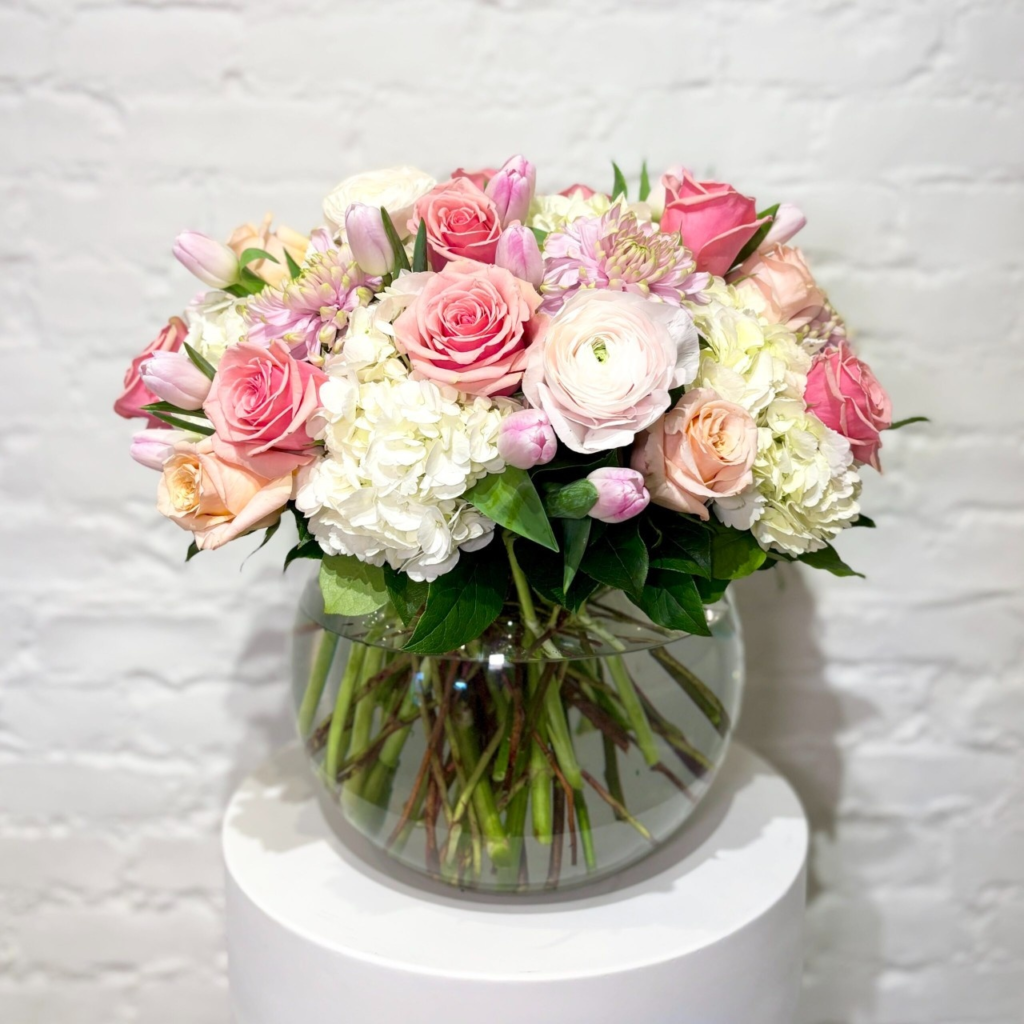 A gorgeous array of spring flowers including ranunculus, roses, and tulips, mixed with fluffy hydrangeas, long-lasting commercial mums and greens in a clear glass bowl.