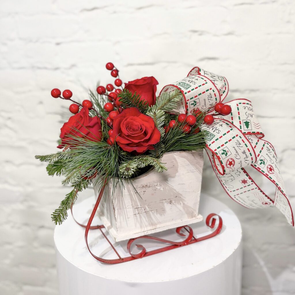 Luscious red roses, red berries and winter greens expertly arranged in a rustic sleigh, accented with a holiday bow.