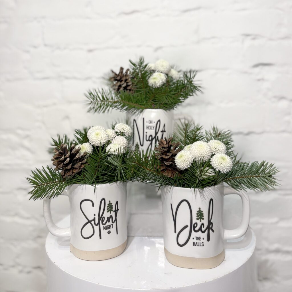 Ceramic mugs with a festive design, filled with fresh winter greens, white poms, and natural pinecones for a soft, snowy woodland look.