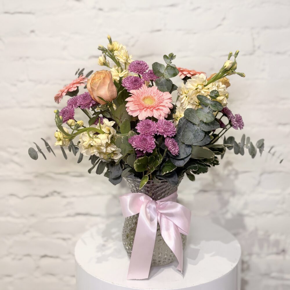 This soft and airy arrangement features a blend of peach and blush roses, cheerful gerbera daisies, delicate stock, textured chrysanthemums, and lush eucalyptus, this bouquet is full of movement and charm. Designed in a classic glass vase finished with a satin pink ribbon, Blossom Breeze feels fresh, feminine, and effortlessly elegant.