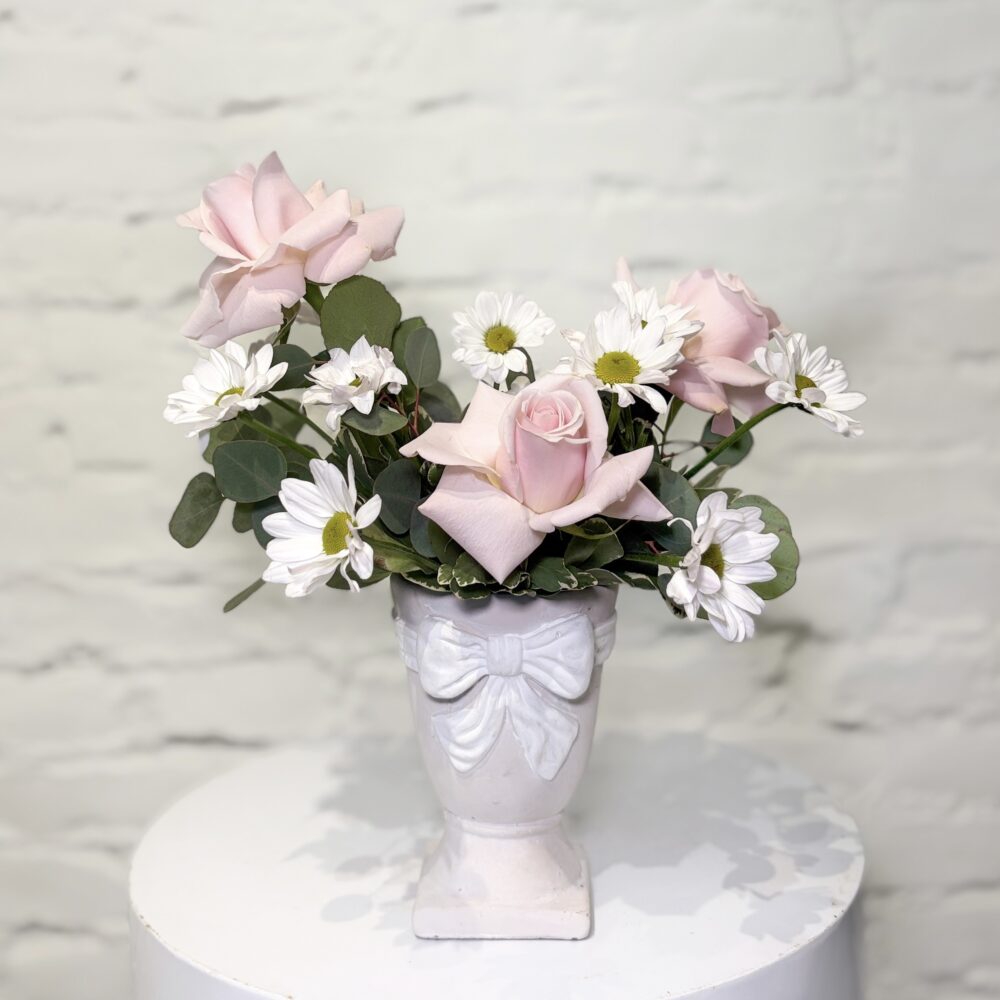Ribboned Grace is a timeless expression of softness and elegance. Featuring delicate blush roses paired with cheerful white daisies and lush eucalyptus greens, this arrangement is designed in a charming pedestal vase adorned with a sculpted bow detail.