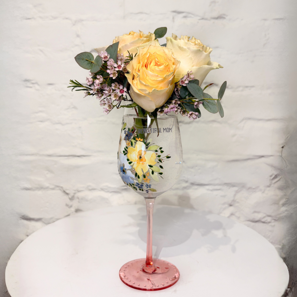 This charming arrangement features soft roses accented with delicate wax flower and fresh greenery, beautifully styled in a keepsake wine glass.