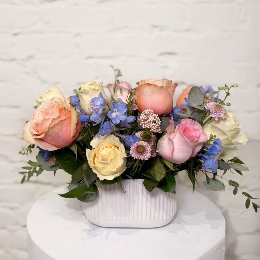 Mixed rose Mother’s Day arrangement with peach, pink, yellow roses and blue delphinium in white vase