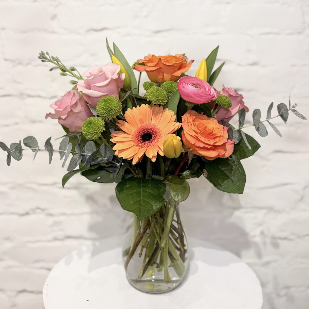 Bright Mother’s Day flower arrangement with peach roses, pink roses, gerbera daisy, ranunculus and tulips in clear vase