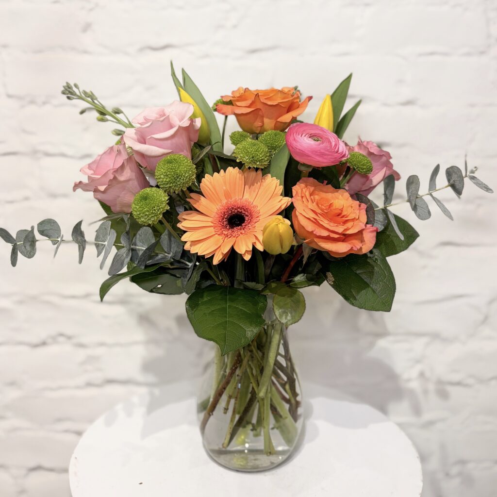 Bright Mother’s Day flower arrangement with peach roses, pink roses, gerbera daisy, ranunculus and tulips in clear vase